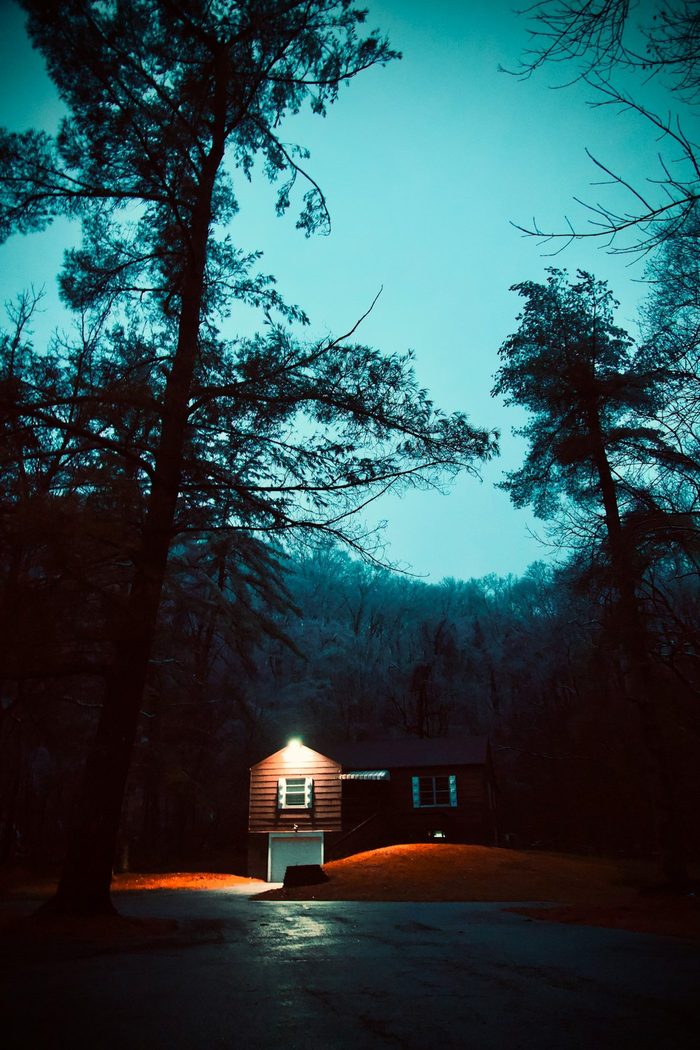 A lone house at night with a single warm lit window, under a teal sky and bare trees